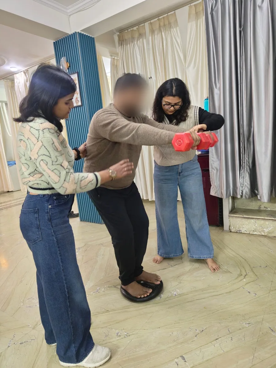 Patient doing supported balance retraining on a wobble board with two physiotherapists at Dynamics Mend Noida